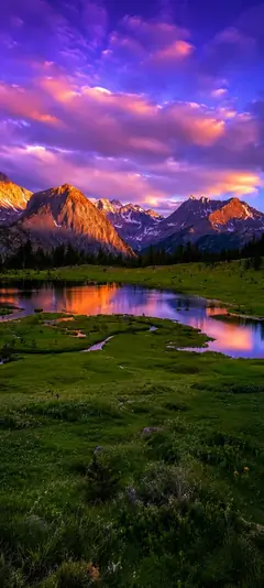 Majestätische Berglandschaft mit ruhigem See, violettem Sonnenuntergang, grüner Wiese und Spiegelung im Wasser als vertikales Handy Wallpaper