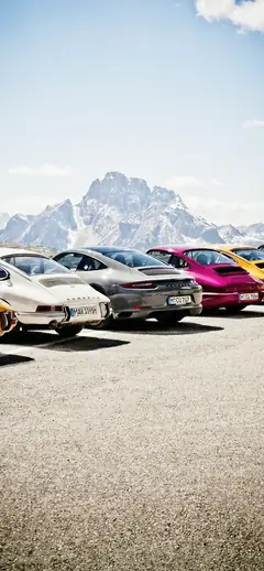 Eine Reihe von klassischen und modernen Porsche 911 Sportwagen geparkt vor einer beeindruckenden, schneebedeckten Berglandschaft als Handy Hintergrundbild.