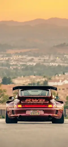 Dunkelroter RWB Porsche 911 von hinten betrachtet, geparkt vor einer Stadtlandschaft und Bergen bei Sonnenuntergang als Handy Hintergrundbild.