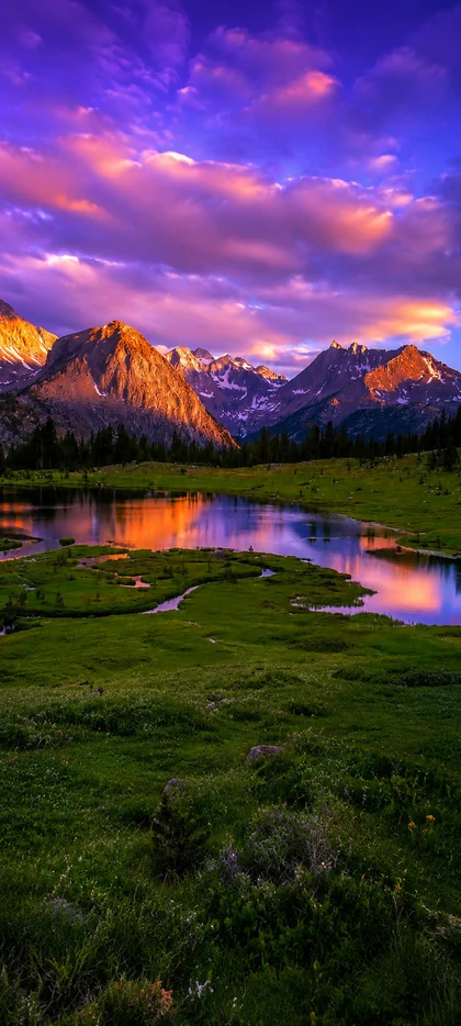 Majestätische Berglandschaft mit ruhigem See, violettem Sonnenuntergang, grüner Wiese und Spiegelung im Wasser als vertikales Handy Wallpaper