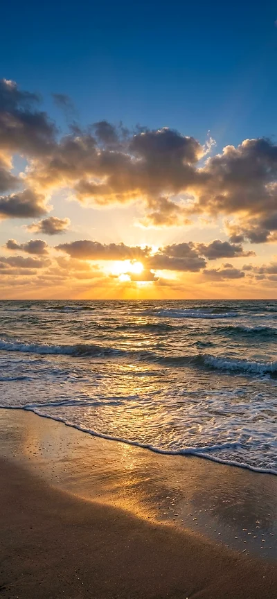 Vertikales Handy Wallpaper: Goldener Sonnenaufgang über dem Ozean mit dramatischen Wolken und Wellen am Sandstrand.