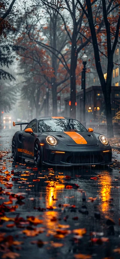 Vertikales iPhone Hintergrundbild: Ein mattschwarzer Porsche 911 GT3 RS mit orangen Rennstreifen und Akzenten parkt auf einer nassen, von Herbstblättern bedeckten Straße im Regen. Die Lichter spiegeln sich auf dem Asphalt.